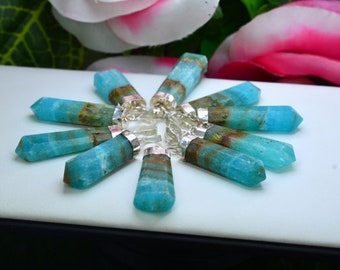 May include: A collection of blue-green gemstone pendants with silver-colored caps, arranged in a circular pattern. The gemstones have a crystalline structure and are displayed on a white surface, with pink and green floral accents in the background.