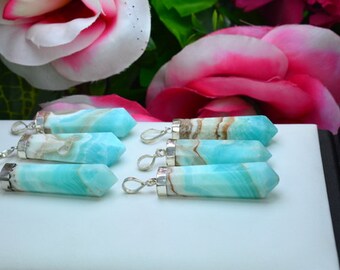 May include: Several blue and white gemstone pendants with silver-colored loops are displayed on a white surface. The pendants are hexagonal in shape and have a polished finish. The background features pink and white flowers.