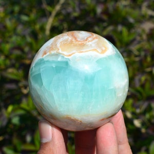 May include: A polished gemstone sphere, predominantly teal and white with areas of tan and brown. The sphere is held in front of a blurred green background. The stone has a smooth, rounded shape, and the colors blend together.