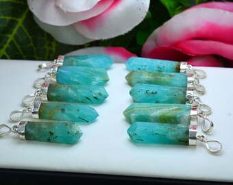 May include: A collection of turquoise-colored crystal pendants with silver-colored metal caps and loops. The crystals have a polished, faceted shape and display natural variations in color. The background features pink and white flowers.