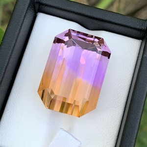 May include: A rectangular gemstone with a gradient of colors, transitioning from a golden-orange at the base to a vibrant purple at the top. The gem is faceted, showcasing its internal structure and clarity, and is set in a black box.