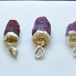 Pode incluir: Sete pingentes de rubi bruto com tampas e laços de metal prateado. As pedras de rubi variam em tamanho e tom, variando do roxo profundo ao marrom avermelhado. Os pingentes são exibidos em uma superfície branca.