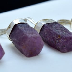 Könnte beinhalten: Vier Silberringe mit jeweils einem rohen, tiefvioletten Edelstein. Die Edelsteine sind zylindrisch mit strukturierter Oberfläche. Die Ringe sind vor einem weißen Hintergrund platziert. Die Steine sind ca. 2-3 cm lang.