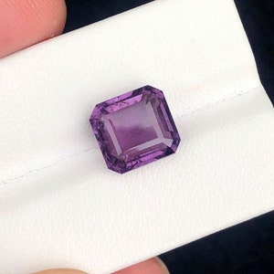 May include: A faceted, rectangular amethyst gemstone with a deep purple hue. The gem is set against a white surface, showcasing its clarity and cut. The gemstone's facets reflect light, highlighting its brilliance. This gemstone is ideal for jewelry making.