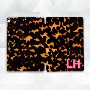 Pode incluir: Capa de tablet aberta com um padrão de tartaruga em tons de castanho e dourado. A capa tem um monograma rosa com as letras "LH". A capa foi concebida para proteger o tablet.