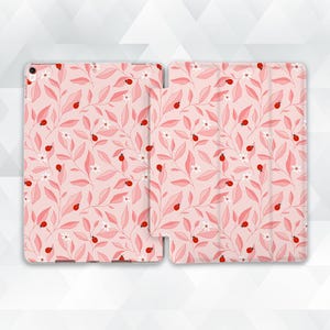 Może przedstawiać: Etui na tablet z powtarzającym się wzorem małych czerwonych biedronek, białych kwiatów i jasnoróżowych liści na delikatnym różowym tle. Wzór pokrywa zarówno tylny panel, jak i składaną przednią okładkę ochronnego etui na urządzenie.