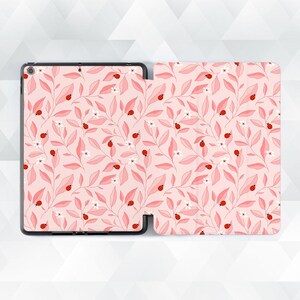 Może przedstawiać: Etui na tablet z powtarzającym się wzorem małych czerwonych biedronek, białych kwiatów i jasnoróżowych liści na delikatnym różowym tle. Etui posiada czarną ramkę oraz składaną okładkę z tym botanicznym motywem i owadami.