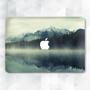 May include: A laptop skin with a misty mountain lake and forest scene. The image features a reflection of the mountains in the water. The Apple logo is visible in the center of the skin.