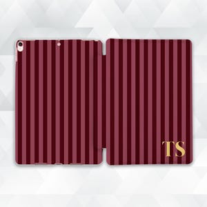 May include: A burgundy and light burgundy striped tablet case, open to display the interior. The case features vertical stripes and gold-colored initials "TS" on the right side. The case is designed to protect a tablet.