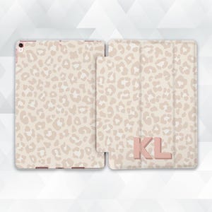 May include: A beige and light brown leopard print tablet case with the initials "KL" in a matching color. The case is open, revealing the tablet inside. The design is repeated on the front and back of the case.