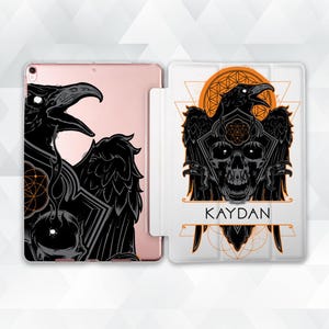 May include: A white iPad case with a black and orange design featuring a skull with two ravens on either side. The text "KAYDAN" is printed below the skull.
