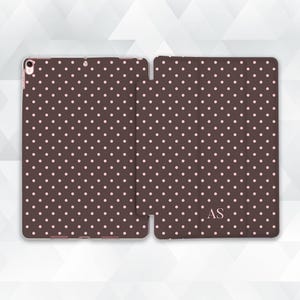 May include: A brown tablet case featuring a pink polka dot design. The case is open, displaying the interior and exterior pattern. The initials "AS" are visible on the cover.