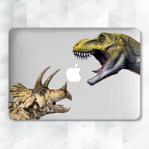 Może przedstawiać: Naklejka na laptopa z szczegółową ilustracją Tyrannosaurus Rex i Triceratops naprzeciwko siebie. Projekt zawiera białe logo Apple. Dinozaury są renderowane w odcieniach zieleni, brązu i beżu na szarym tle.