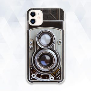 May include: A clear phone case featuring a vintage camera design. The case has a black and silver camera graphic with two lenses and a dial. The phone's camera lenses are visible at the top.