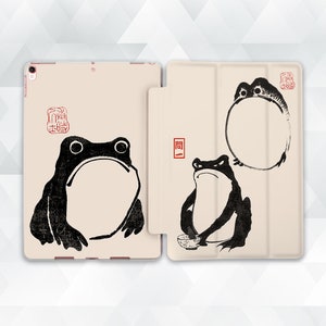 May include: A beige iPad case with a black and white illustration of three frogs. The case has a flap that covers the screen. The frogs are in different poses, one is sitting, one is standing, and one is in a circle.