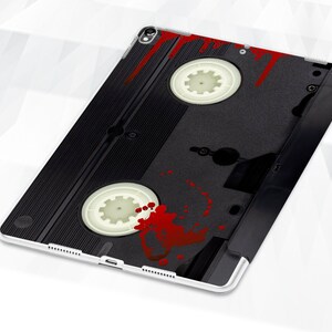 May include: A black and white VHS cassette tape with red blood splatter on the front. The tape is on a white background.
