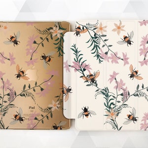 以下が含まれることがあります： 蜂と葉が描かれた花柄の金のiPadケース。ケースは白地にピンクと緑のアクセントが施されています。iPadケースはiPadに合うように設計されています。