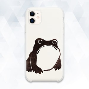 May include: A clear phone case with a brown frog illustration. The frog has large eyes and a wide open mouth. The design is printed on a white background. The phone case is designed to fit a smartphone.