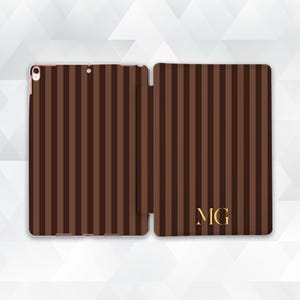 May include: A tablet case featuring a vertical striped pattern in alternating shades of dark and medium brown. The right side of the case displays the gold-colored initials MG in a serif font. The case includes a camera cutout on the left side.
