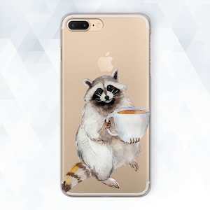 Puede incluir: Una funda de teléfono transparente con un borde dorado y una ilustración de acuarela de un mapache sosteniendo una taza de café. El mapache es gris y blanco con ojos negros y una nariz negra.