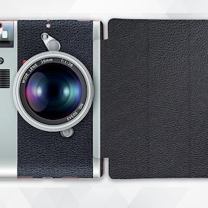 May include: A black and silver camera case with a faux leather texture. The front of the case features a vintage camera design with a lens, a flash, and the text "WIDE LENS 35mm f:1:28 EPEOS ON".