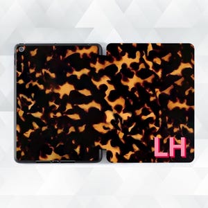 Può includere: Una custodia per tablet aperta con un motivo a scaglie di tartaruga nei toni del marrone e del giallo. Il lato destro della custodia presenta le lettere rosa "LH". La custodia del tablet è nera.