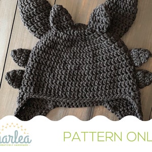 Puede incluir: Sombrero de crochet negro con diseño de dragón. El sombrero tiene dos orejas grandes y un hocico largo. El sombrero está hecho con un punto texturizado. El texto "PATTERN ONLY" es visible en la parte inferior de la imagen.
