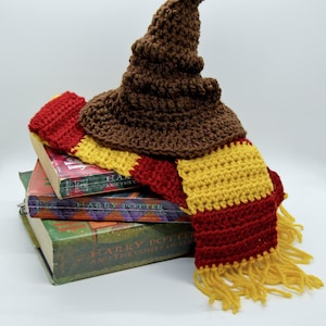 May include: A brown crocheted wizard hat with a pointed top, sitting on top of a stack of three Harry Potter books. A red and gold striped scarf with tassels is draped over the books and hat.