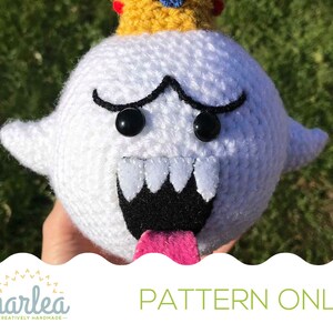 May include: Crochet pattern for a King Boo plush toy. The plush is white with black eyes, a black mouth with white teeth, and a pink tongue sticking out. The plush is wearing a yellow crown with blue and red accents.