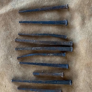 May include: A collection of antique, dark metal nails, each with a rectangular head and a tapered, pointed end. The nails are arranged on a textured, tan fabric surface, showcasing their aged appearance and historical significance.