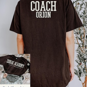 May include: Black t-shirt with white text on the back that reads "COACH ORION". A smaller image shows a black t-shirt with white text that reads "ZODIAC ACADEMY VIPITBALL". The text "Dark Chocolate" is written below the smaller image.