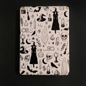 May include: A cream-colored case featuring a repeating black pattern of witch-themed illustrations. The design includes witches, bats, potion bottles, candles, ghosts, and skulls. The case is rectangular with rounded corners.