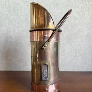 May include: A vintage brass coal scuttle with a copper band and handle. The scuttle has a curved top and a rectangular detail on the side. The handle is made of a silver-coloured metal. The scuttle is cylindrical in shape.