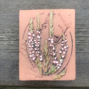 May include: A terracotta tile featuring a raised design of pink heather flowers and green leaves.