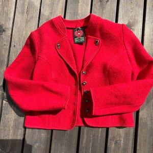 Puede incluir: Una chaqueta de lana roja con cuello alto, cierre de botones y costuras decorativas. La chaqueta tiene una peque&ntilde;a etiqueta con un logotipo de p&aacute;jaro.