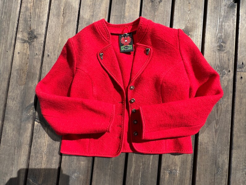 Puede incluir: Una chaqueta de lana roja con cuello alto, cierre de botones y costuras decorativas. La chaqueta tiene una peque&ntilde;a etiqueta con un logotipo de p&aacute;jaro.