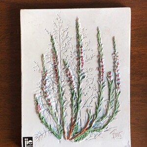 May include: A white ceramic tile featuring a raised relief design of a cluster of green and red heather flowers. The tile is signed "Thuis" in the bottom right corner and has a "JIE" logo in the bottom left corner.