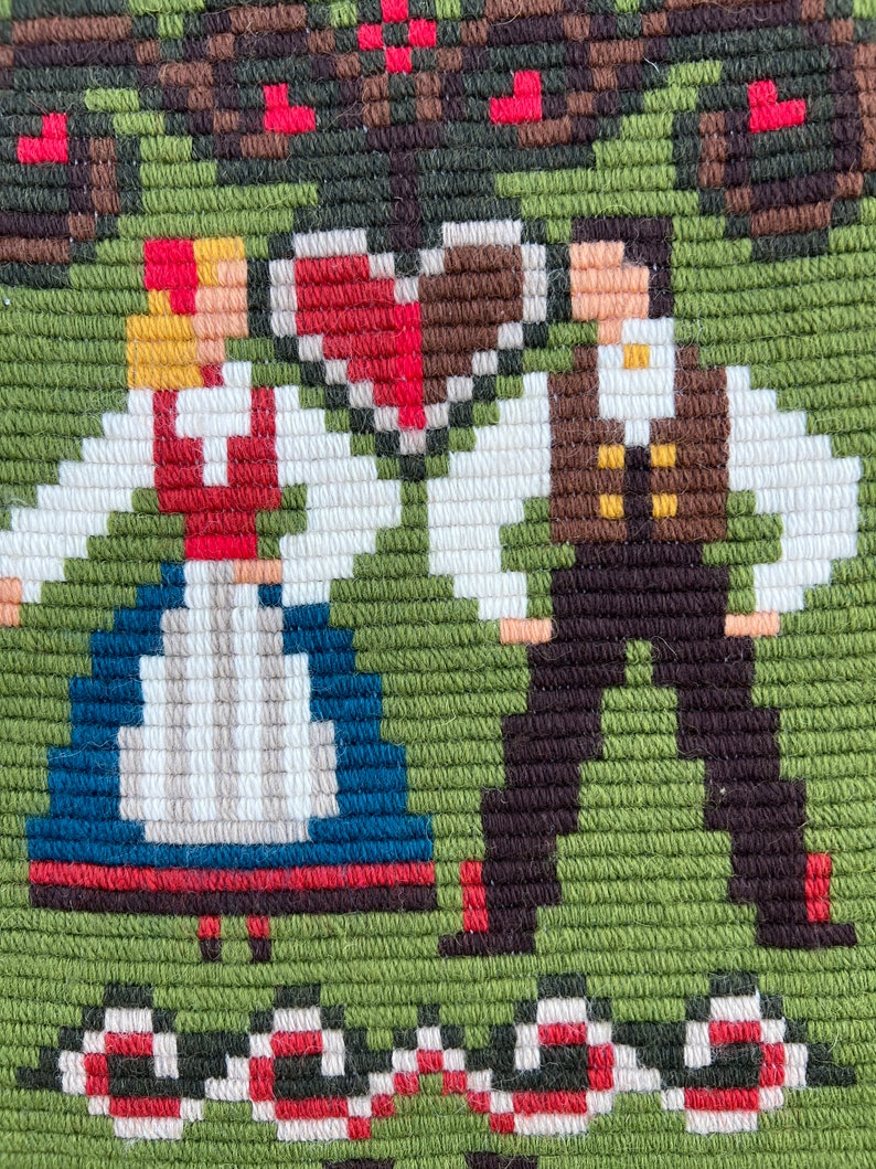 May include: Embroidered wall hanging featuring a couple in traditional folk costumes, standing under a heart-shaped design with red and brown accents. The background is green with a red and white border.