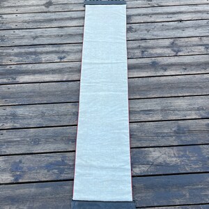 May include: A long, white fabric scroll hanging from a black metal scroll holder with decorative details. The scroll is hanging against a wooden background.