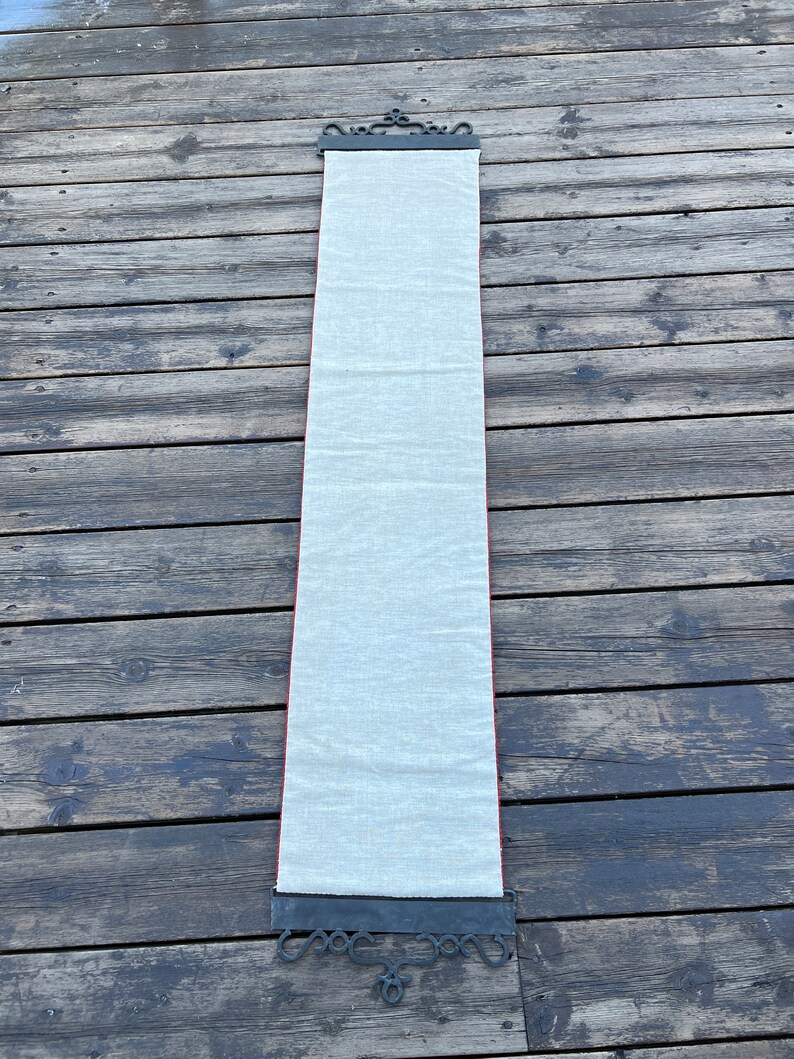 May include: A long, white fabric scroll hanging from a black metal scroll holder with decorative details. The scroll is hanging against a wooden background.