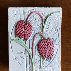 May include: Square ceramic tile with a raised floral design. The tile features two pink and red checkered flower heads with green stems and leaves against a light blue background. The background has a textured, embossed pattern.
