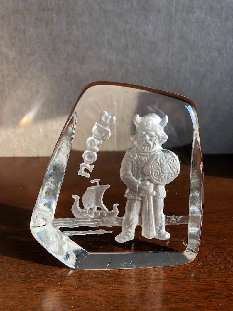 May include: A clear glass paperweight with a white etched design of a Viking warrior holding a shield and sword. The word "Norse" is etched on the side of the paperweight.