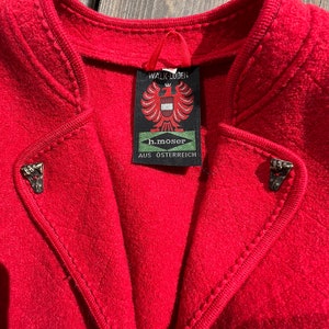 Puede incluir: Una chaqueta de lana roja con una etiqueta que dice "Walk Loden h.moser AUS OSTERREICH". La chaqueta tiene un cuello alto y costuras decorativas alrededor de los bordes.