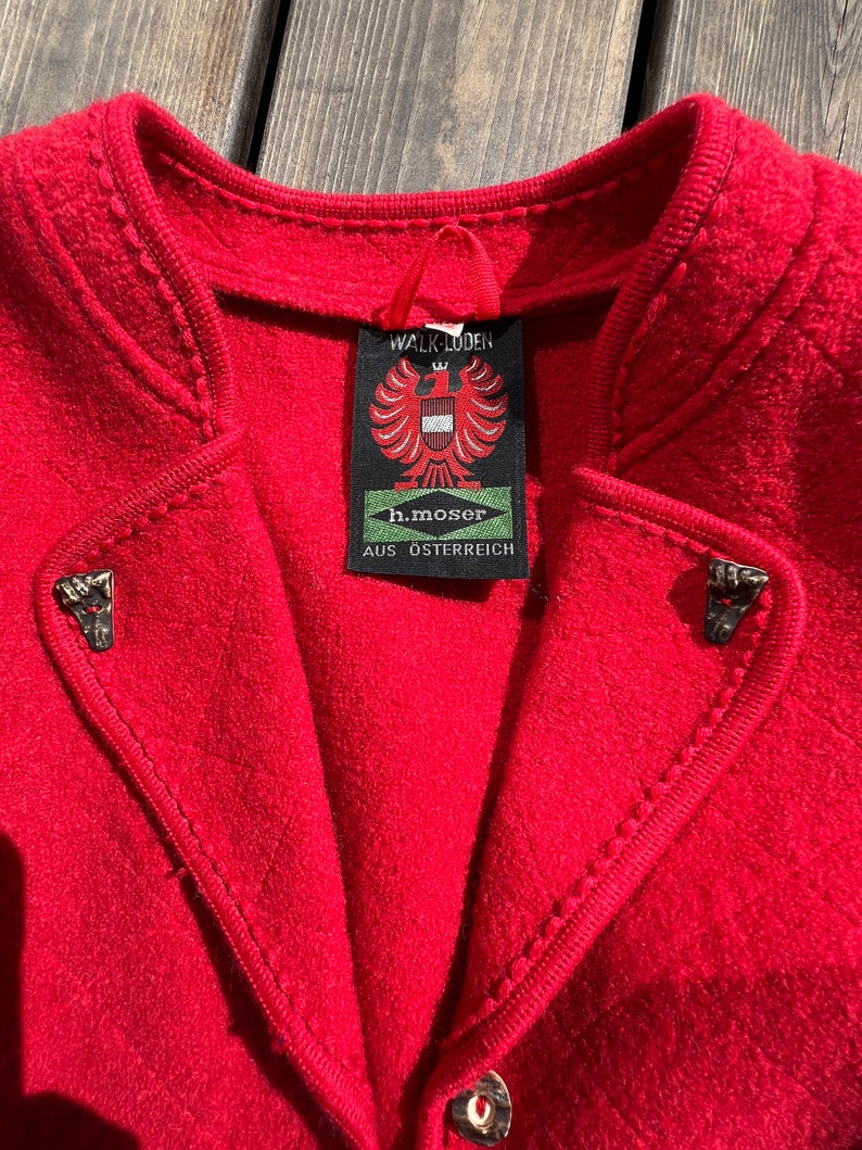 Puede incluir: Una chaqueta de lana roja con una etiqueta que dice "Walk Loden h.moser AUS OSTERREICH". La chaqueta tiene un cuello alto y costuras decorativas alrededor de los bordes.