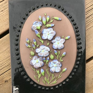 May include: A brown ceramic tile with a black frame and a raised oval design featuring blue and green flowers. The tile is signed "JIE GANTORPTA SWEDEN".