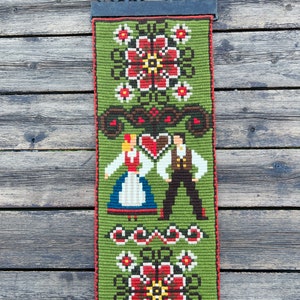 May include: A green tapestry with a traditional folk design featuring a couple in traditional clothing, flowers, and geometric patterns. The tapestry is hanging on a wooden wall.