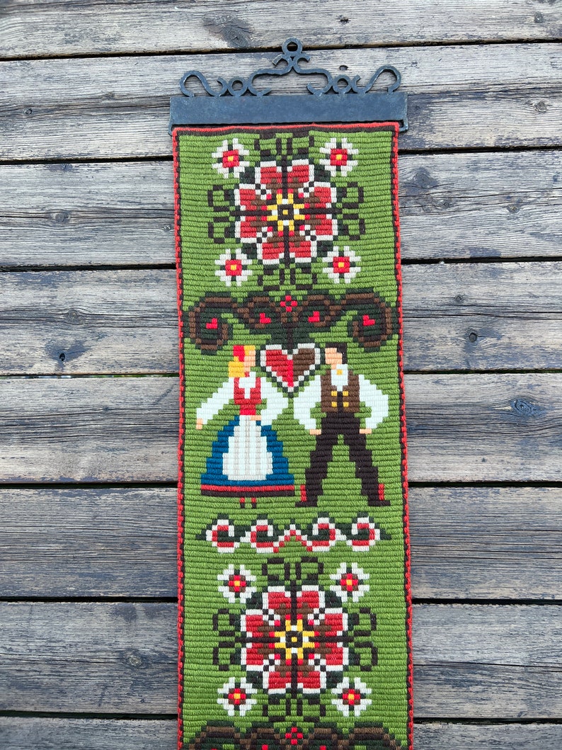 May include: A green tapestry with a traditional folk design featuring a couple in traditional clothing, flowers, and geometric patterns. The tapestry is hanging on a wooden wall.