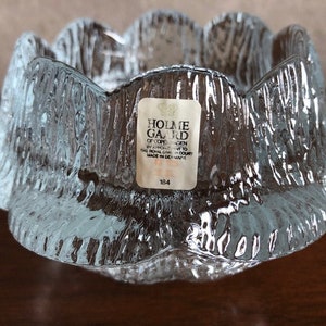 May include: A clear glass candle holder with a textured surface. The holder has a scalloped edge and is made by Holme Gard of Copenhagen, Denmark. The label on the holder says "Holme Gard of Copenhagen by appointment to the Royal Danish Court Made in Denmark 134".
