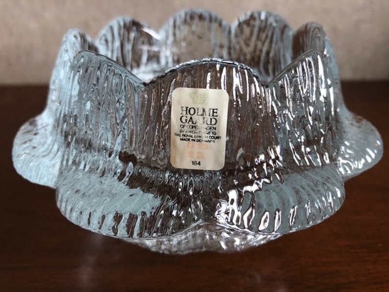 May include: A clear glass candle holder with a textured surface. The holder has a scalloped edge and is made by Holme Gard of Copenhagen, Denmark. The label on the holder says "Holme Gard of Copenhagen by appointment to the Royal Danish Court Made in Denmark 134".
