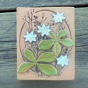May include: A terracotta-coloured tile with a raised relief of a floral design. The design features white flowers, green leaves, and delicate stems within a circular frame. The tile has a rustic, handcrafted appearance.